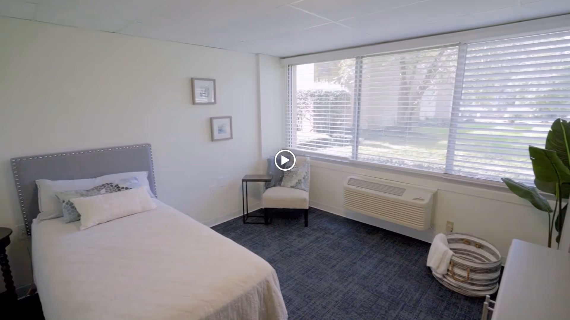 A bright and clean senior living bedroom with a single bed featuring white bedding and decorative pillows. The room has a gray upholstered headboard, a small side table, a cushioned chair near a large window with blinds, a wall-mounted air conditioning unit, a basket with a towel, and a potted plant in the corner.