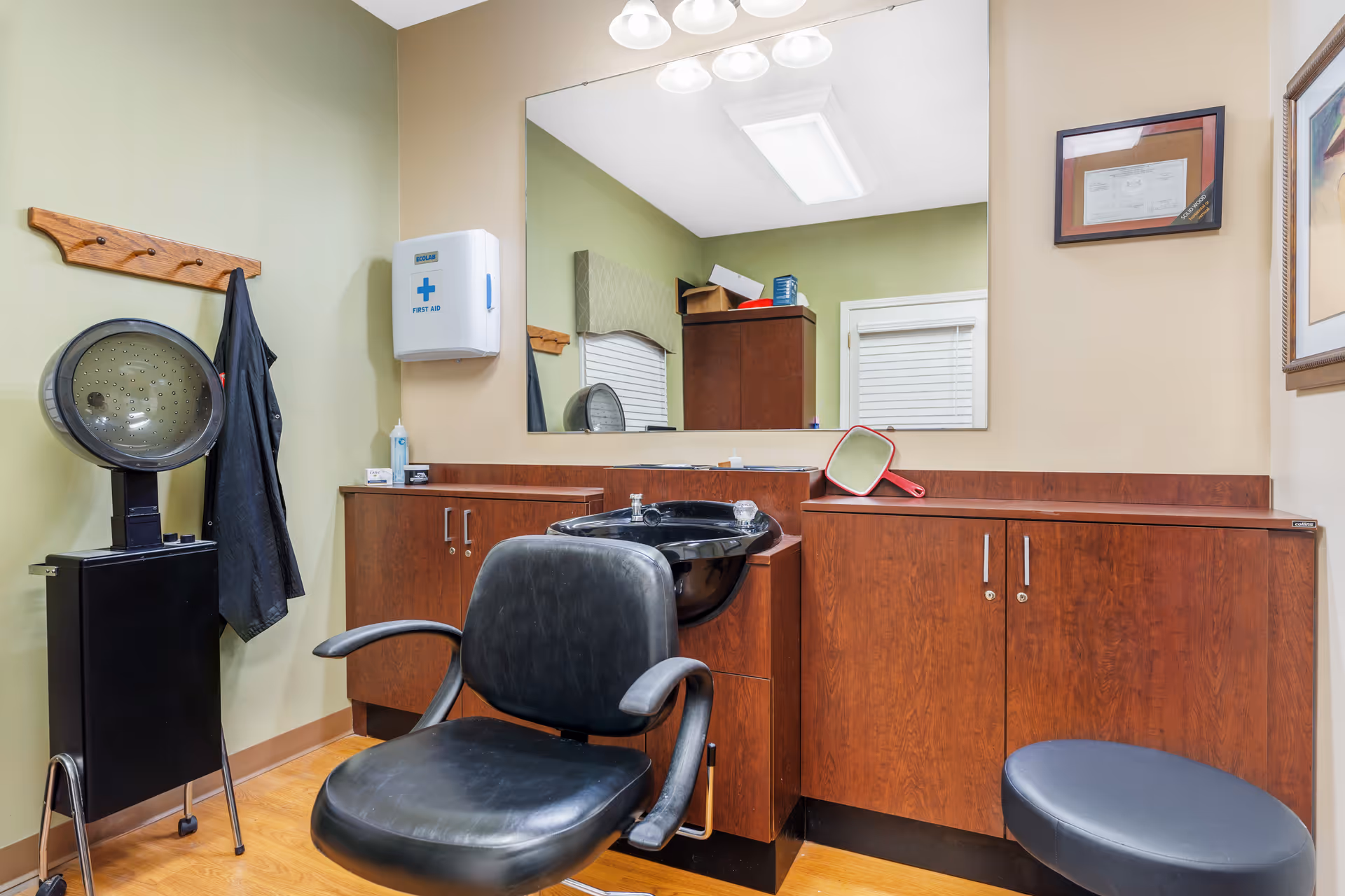 Interior salon room with a black styling chair and wash station, hooded dryer, large mirror, and wood cabinets.