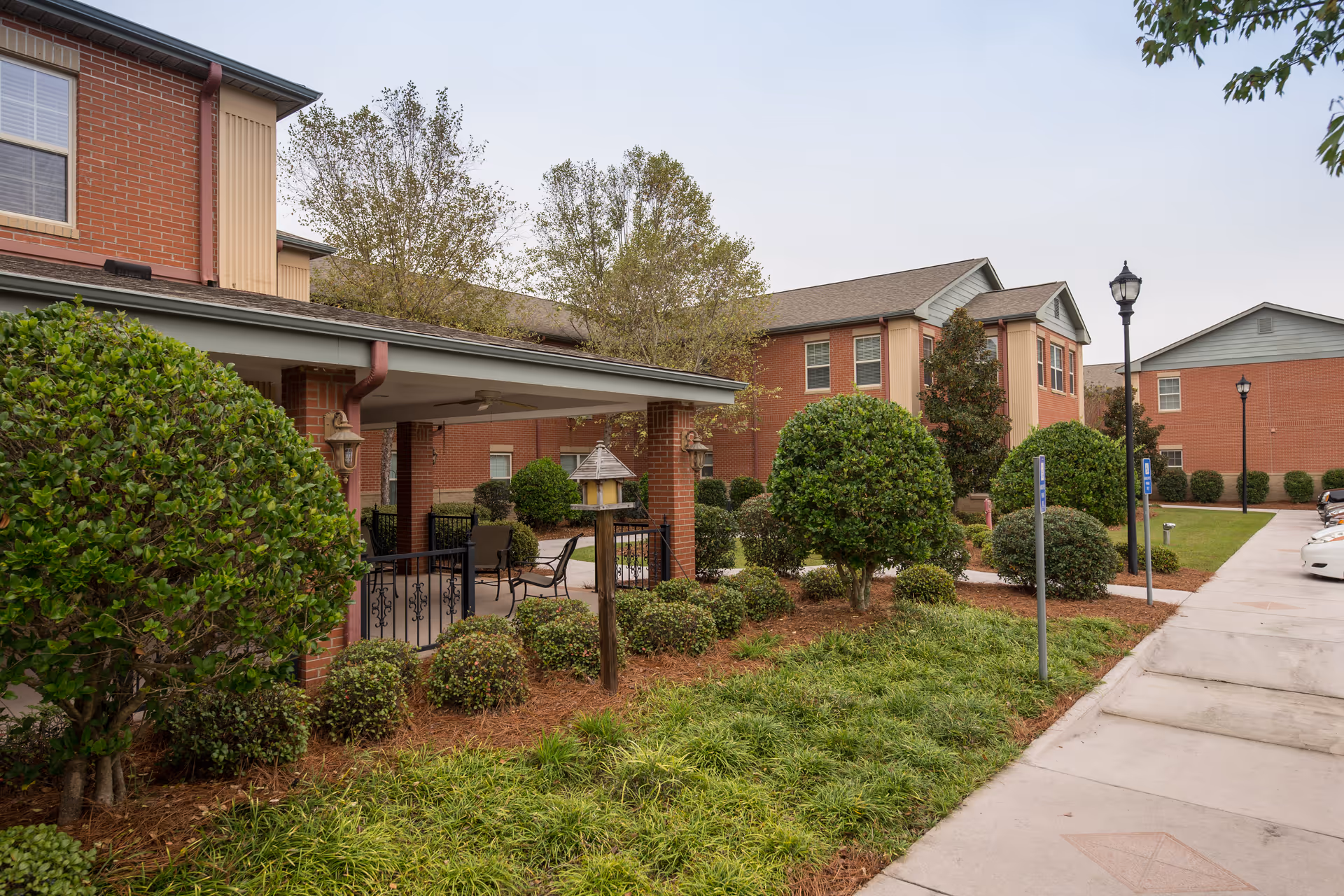 Exterior view of Crescent Landing at Hattiesburg showing a brick building with multiple windows, a covered patio area with outdoor seating, neatly trimmed bushes, trees, and a sidewalk leading to a parking area with cars.