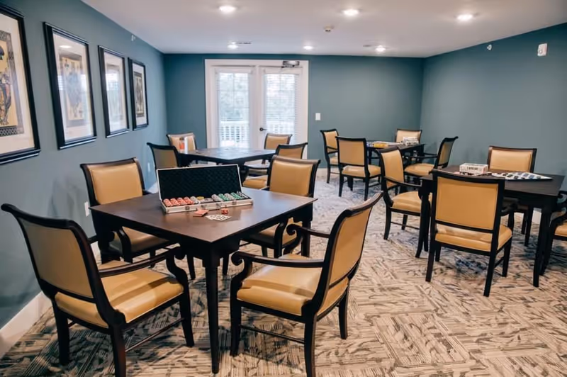 Well-lit common room with several dark wood tables and tan upholstered chairs, some tables set up with games and poker chips.