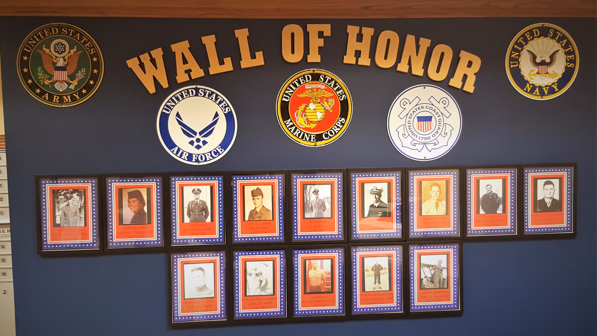 A Wall of Honor display featuring framed photographs of military personnel on a dark blue wall. Above the photos are large emblems and text representing the United States Army, Air Force, Marine Corps, Coast Guard, and Navy. The words 'WALL OF HONOR' are prominently displayed in large yellow letters above the emblems and photos.