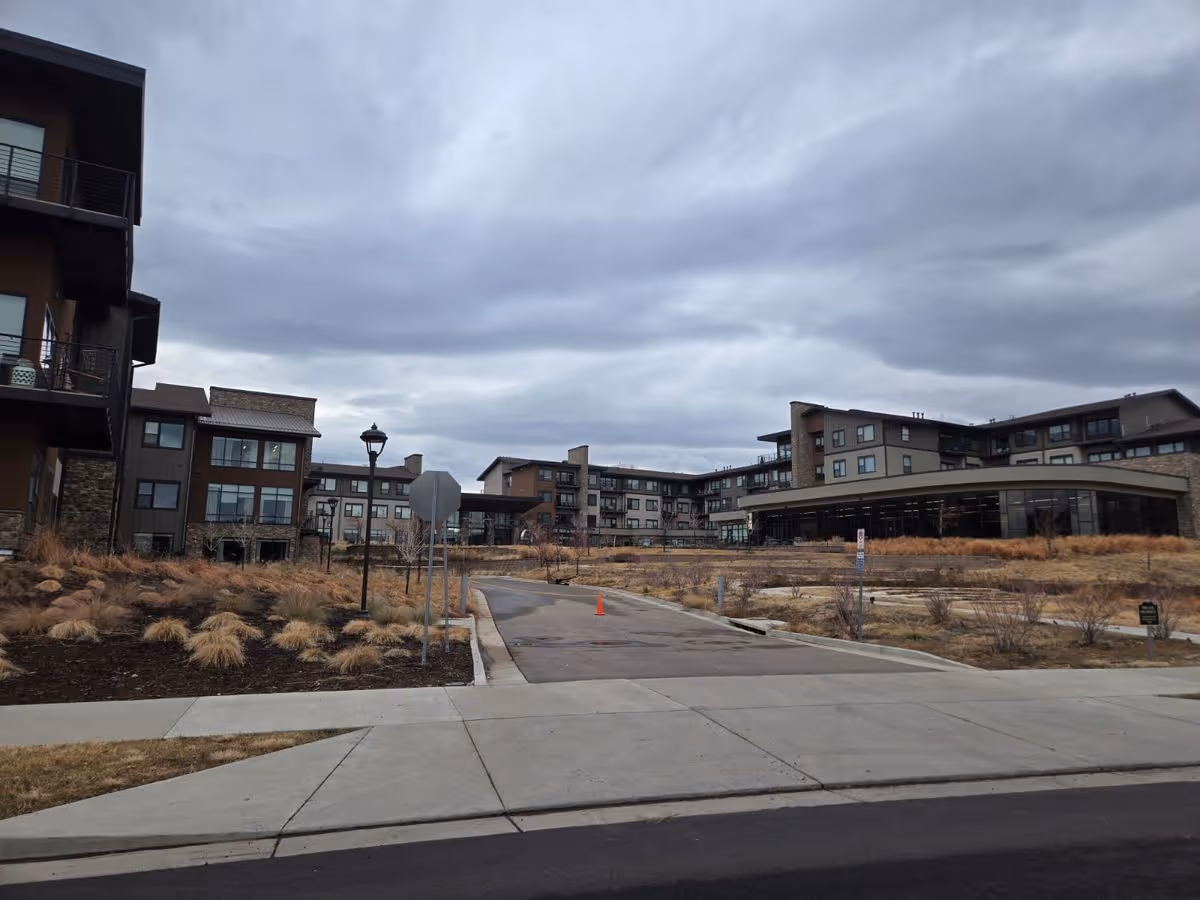 Exterior view of a multi-story senior living facility with a driveway, landscaped grounds, and cloudy sky.