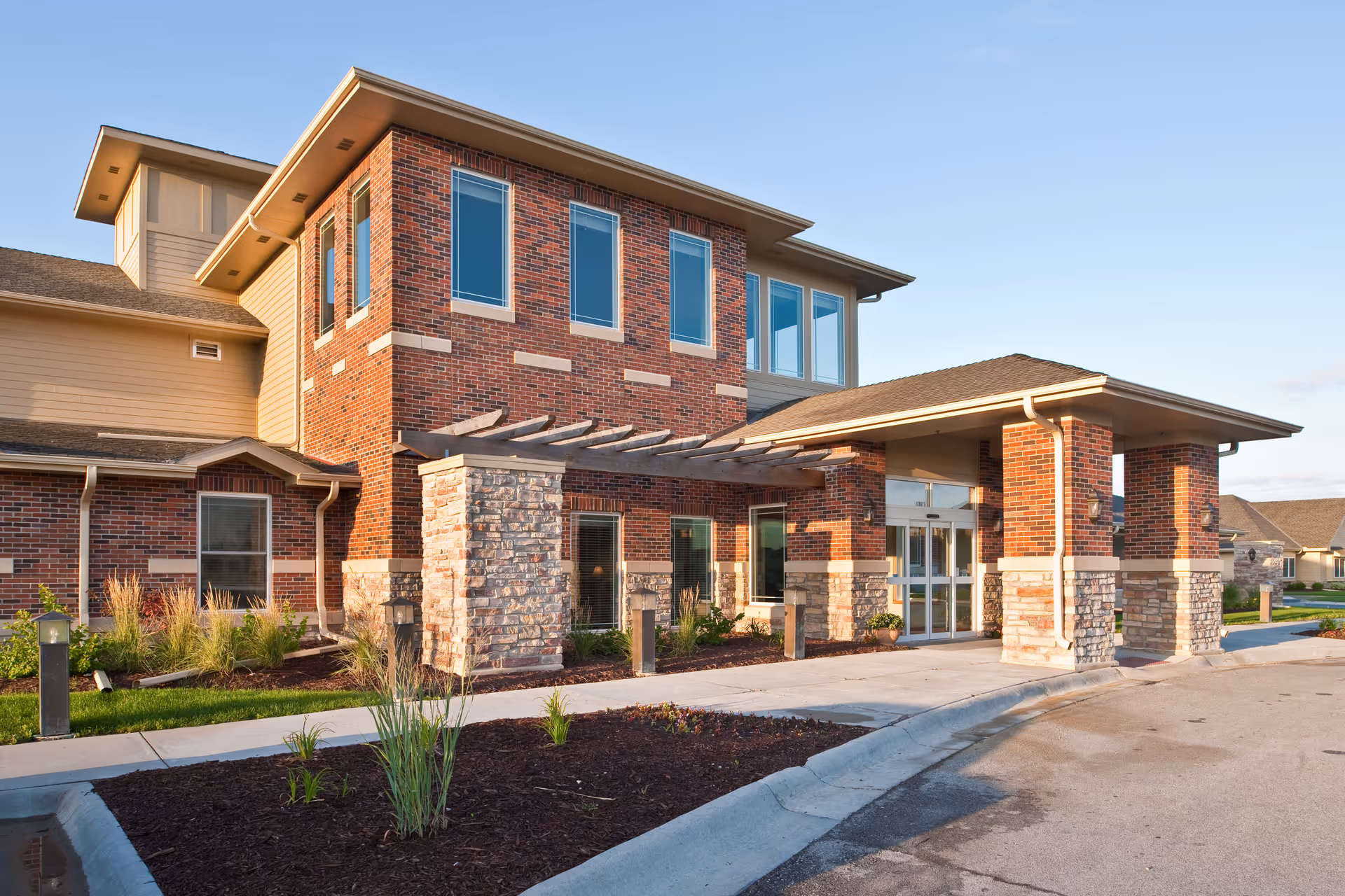 Exterior view of a senior living facility building with brick and stone facade, large windows, and a covered entrance with a pergola. The surrounding area includes landscaped plants and a paved driveway.
