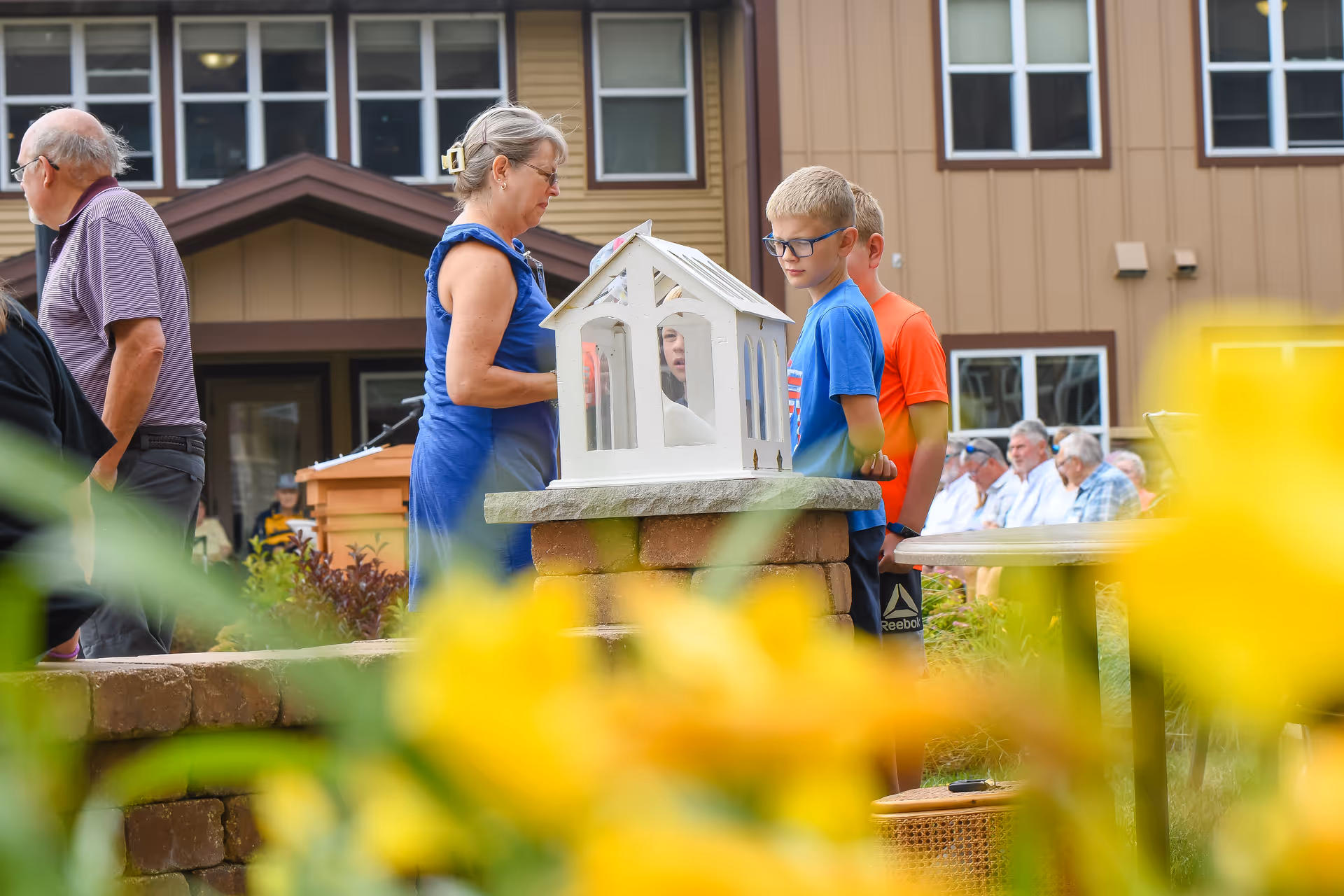 People, including children and seniors, gathered outdoors near a small white decorative house in a courtyard with yellow flowers in the foreground and a building in the background.