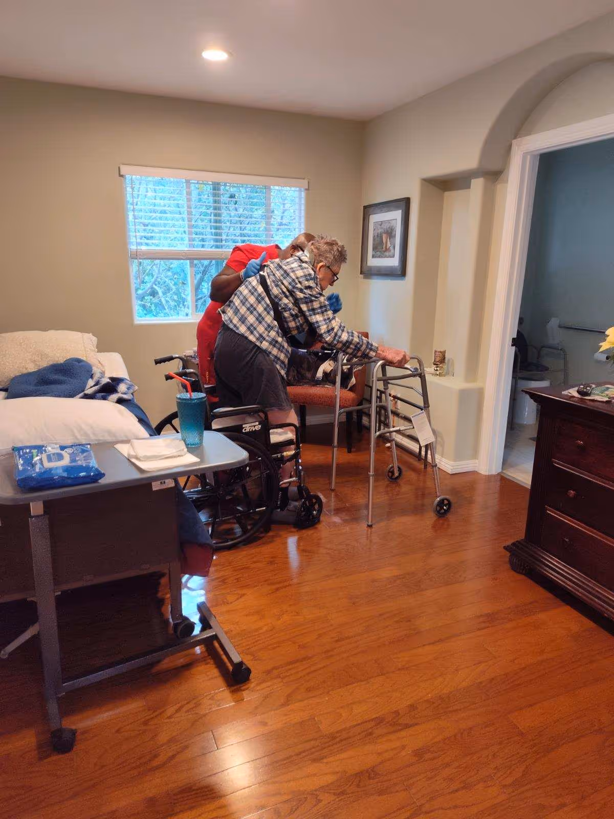 An elderly person using a walker is assisted by a caregiver in a room with wooden floors. The room contains a hospital-style bed with a blue blanket and a small table with a cup and tissues. There is a window with blinds, a framed picture on the wall, and an open doorway leading to a bathroom.