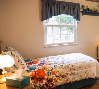 A bedroom with a single bed covered in a floral patterned bedspread. There is a window with a dark curtain valance above the bed, and a wooden nightstand with a lamp and a box of tissues next to the bed. A small shelf with decorative items is mounted on the wall above the bed.