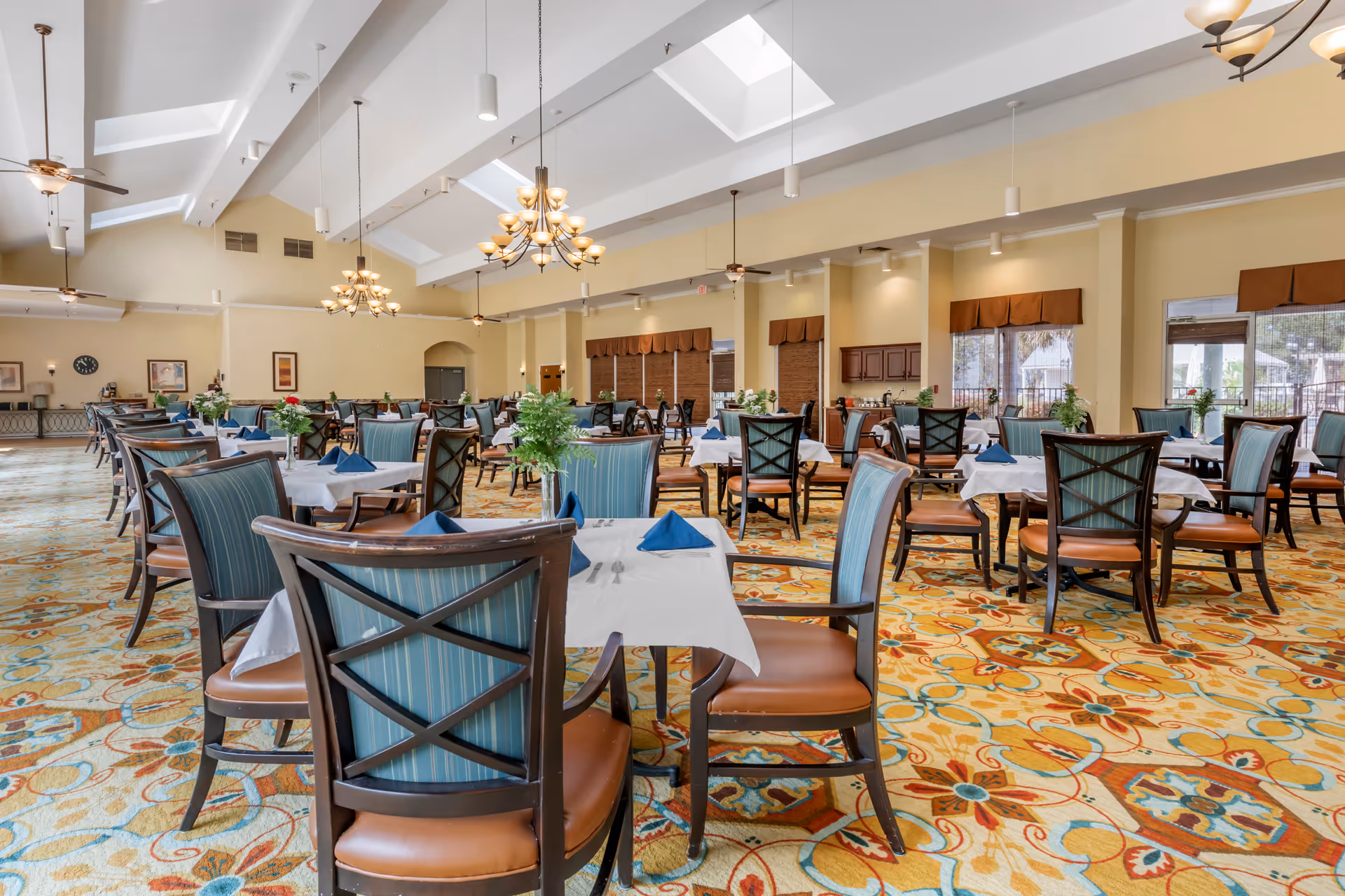 A spacious dining room with multiple tables covered in white tablecloths and set with blue folded napkins and silverware. The chairs have wooden frames with blue striped upholstery and brown leather seats. The room features a colorful patterned carpet, high ceilings with skylights, ceiling fans, and chandeliers. Large windows with brown valances let in natural light.