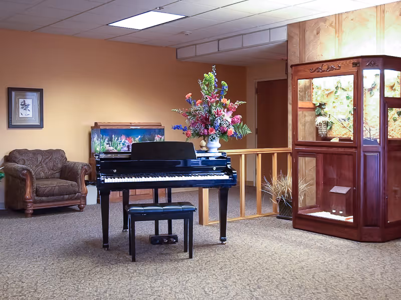 A cozy common area with a black upright piano and matching bench in the center. Behind the piano is a large floral arrangement in a white vase. To the left, there is a brown upholstered armchair and a fish tank on a wooden stand. On the right side, there is a wooden display cabinet with glass panels showcasing decorative items. The walls are painted a warm beige color and the floor is carpeted.