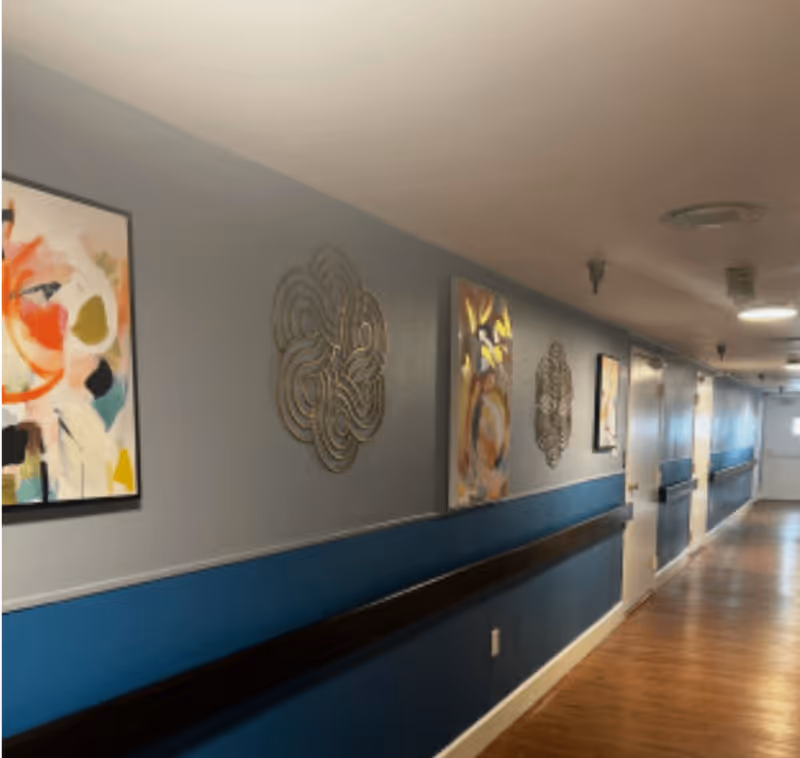 A long indoor hallway with wooden flooring and blue and gray walls. The walls are decorated with abstract paintings and metal wall art. Several white doors line the hallway, and ceiling lights illuminate the space.