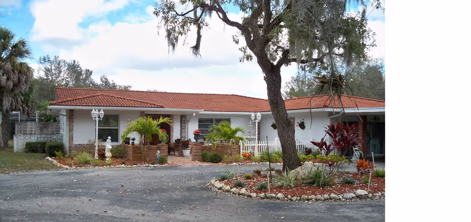 Single-story white residential building with a red tile roof, landscaped front yard, and circular driveway under a large tree.