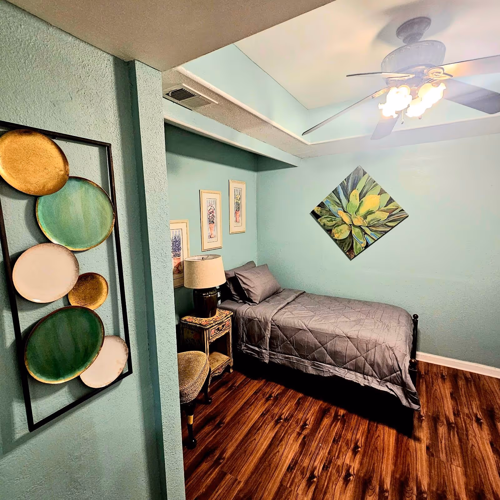 A small bedroom with teal walls and wood flooring featuring a single bed with gray bedding. There is a ceiling fan with lights above the bed. On the wall above the bed is a diamond-shaped painting of green and yellow leaves. Next to the bed is a small decorative nightstand with a lamp and a cushioned chair. On the left wall, there is a decorative wall hanging with circular plates in green, white, and gold colors.