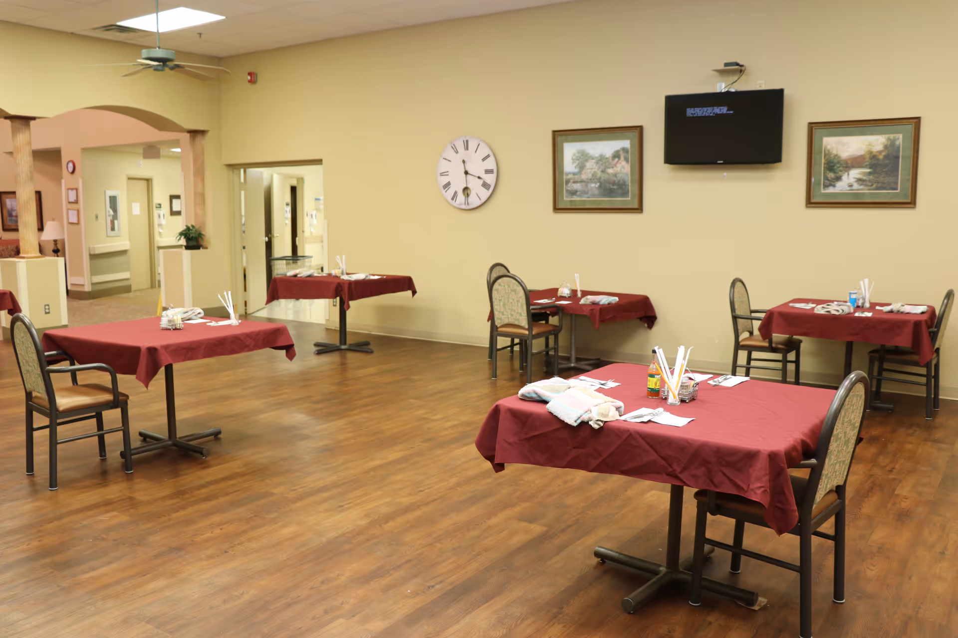 A dining room with four tables covered in red tablecloths, each set with utensils and napkins. The room has wooden flooring, beige walls, a wall clock, two framed paintings, and a wall-mounted television. There are chairs around the tables and an open doorway leading to another area.