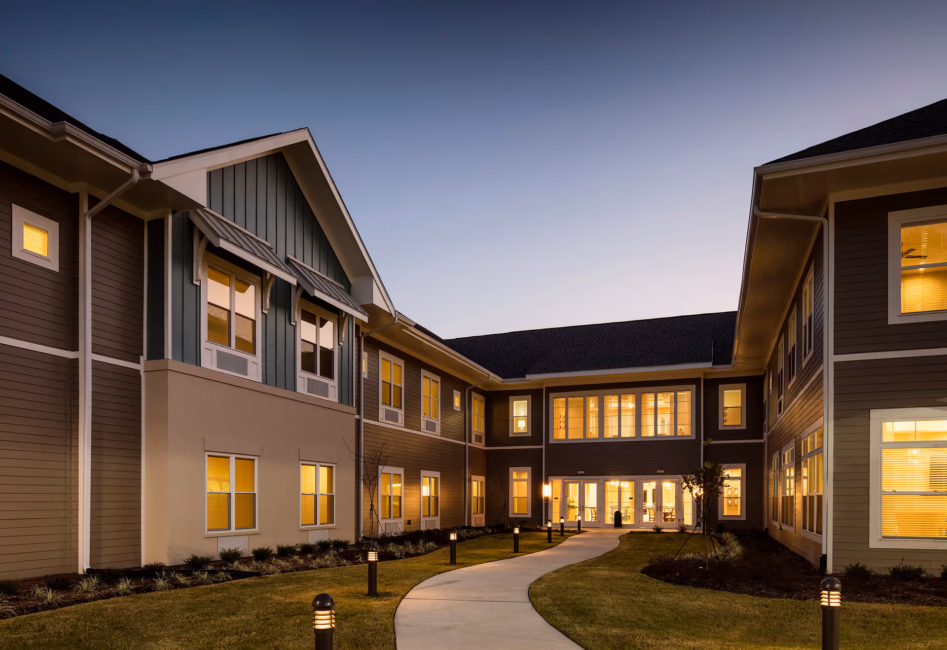 Exterior view of a senior living facility building at dusk with warm lights glowing from the windows. A curved walkway with small lamp posts leads through a landscaped lawn towards the entrance.
