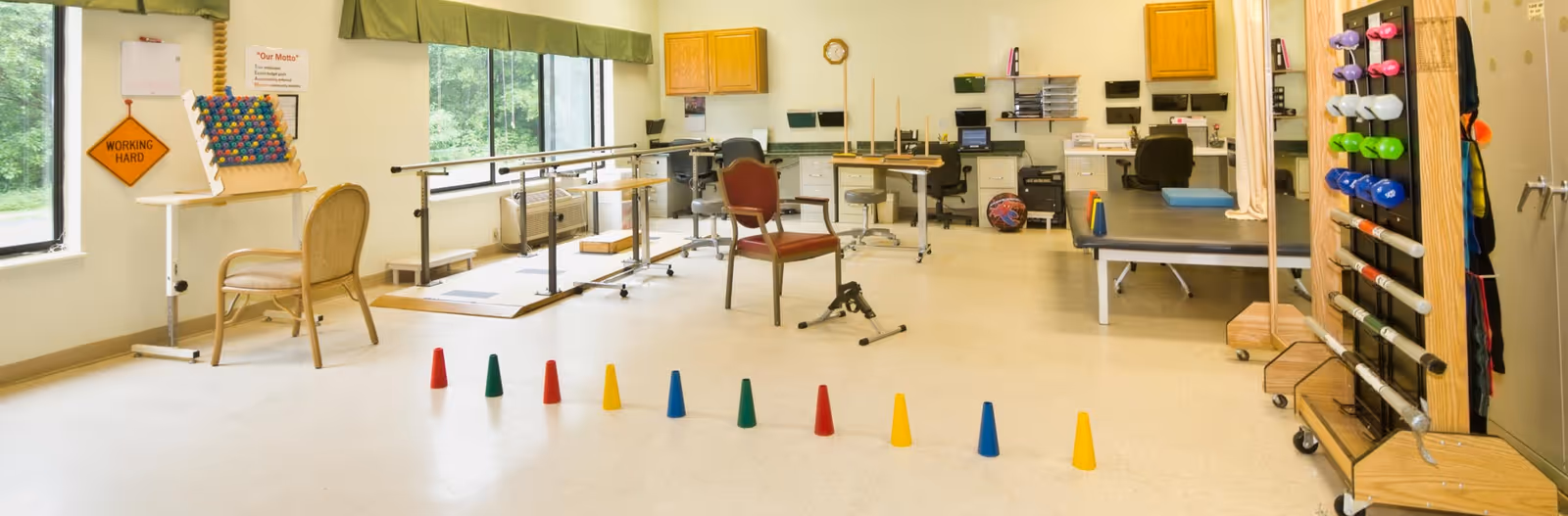 A bright therapy room with large windows, parallel bars for walking exercises, chairs, exercise equipment, colorful cones arranged on the floor, and a rack holding various weights and therapy tools.