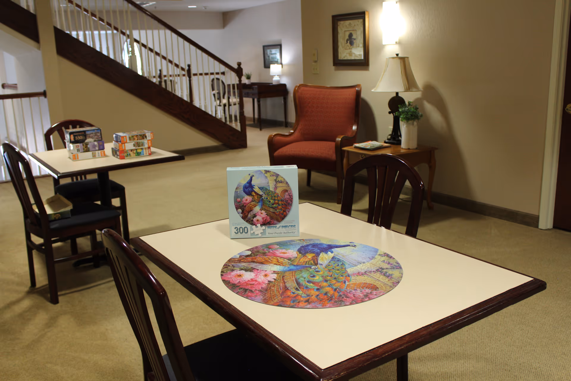 A senior living common area with tables and chairs, a colorful round jigsaw puzzle on a table, and a red armchair in the background.