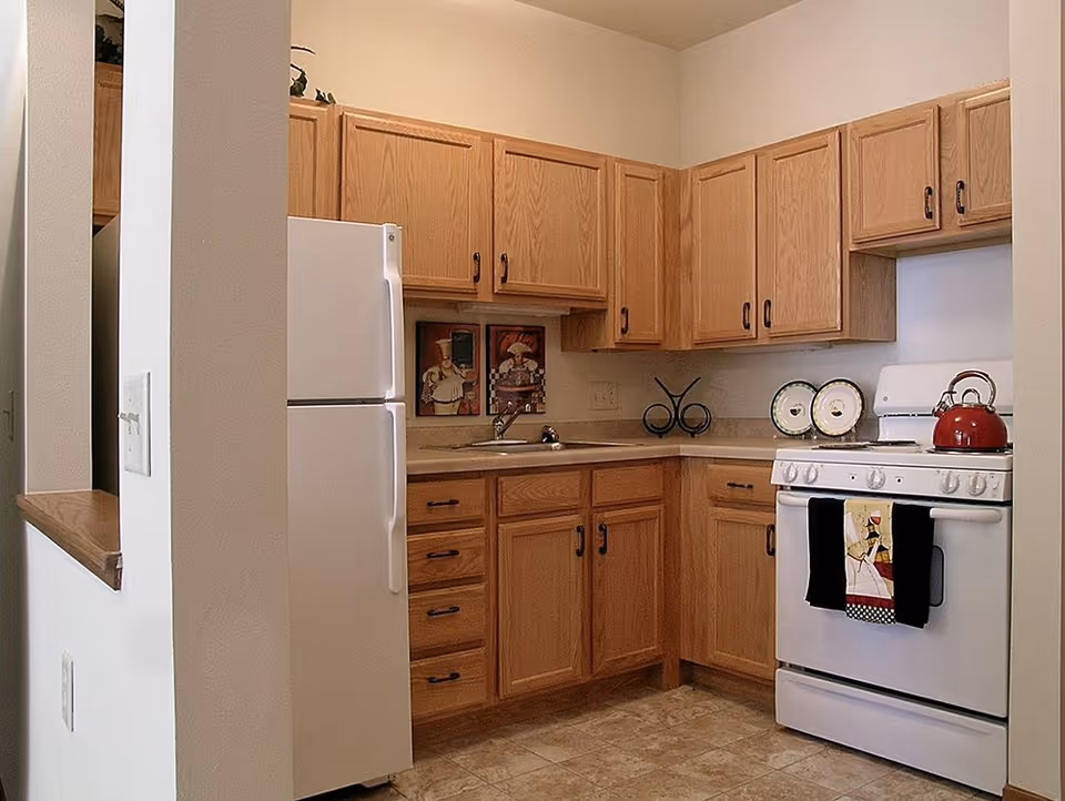 Small kitchen with oak cabinets, a white refrigerator and stove, tiled floor, and a red kettle on the stove.