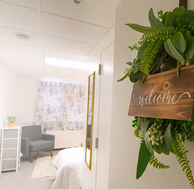 View into a cozy senior living bedroom with a gray armchair, a small white dresser with a potted plant, a bed with white bedding, and floral curtains. A decorative green wreath with a wooden sign that says 'welcome' hangs on the wall near the door.