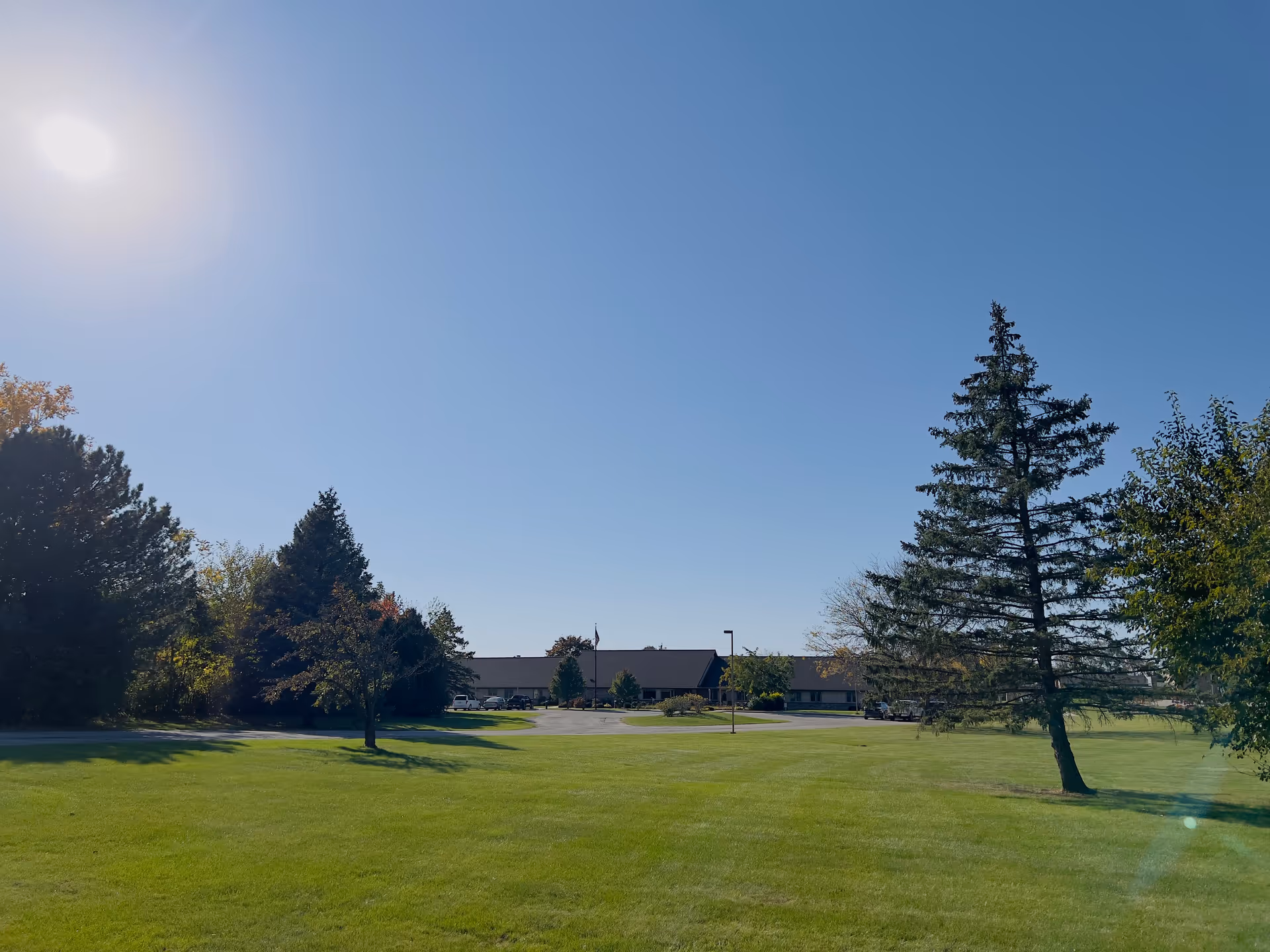 A wide view of a green lawn with scattered trees under a clear blue sky. In the distance, there is a single-story building with a parking lot and several cars parked. The sun is shining brightly in the sky.