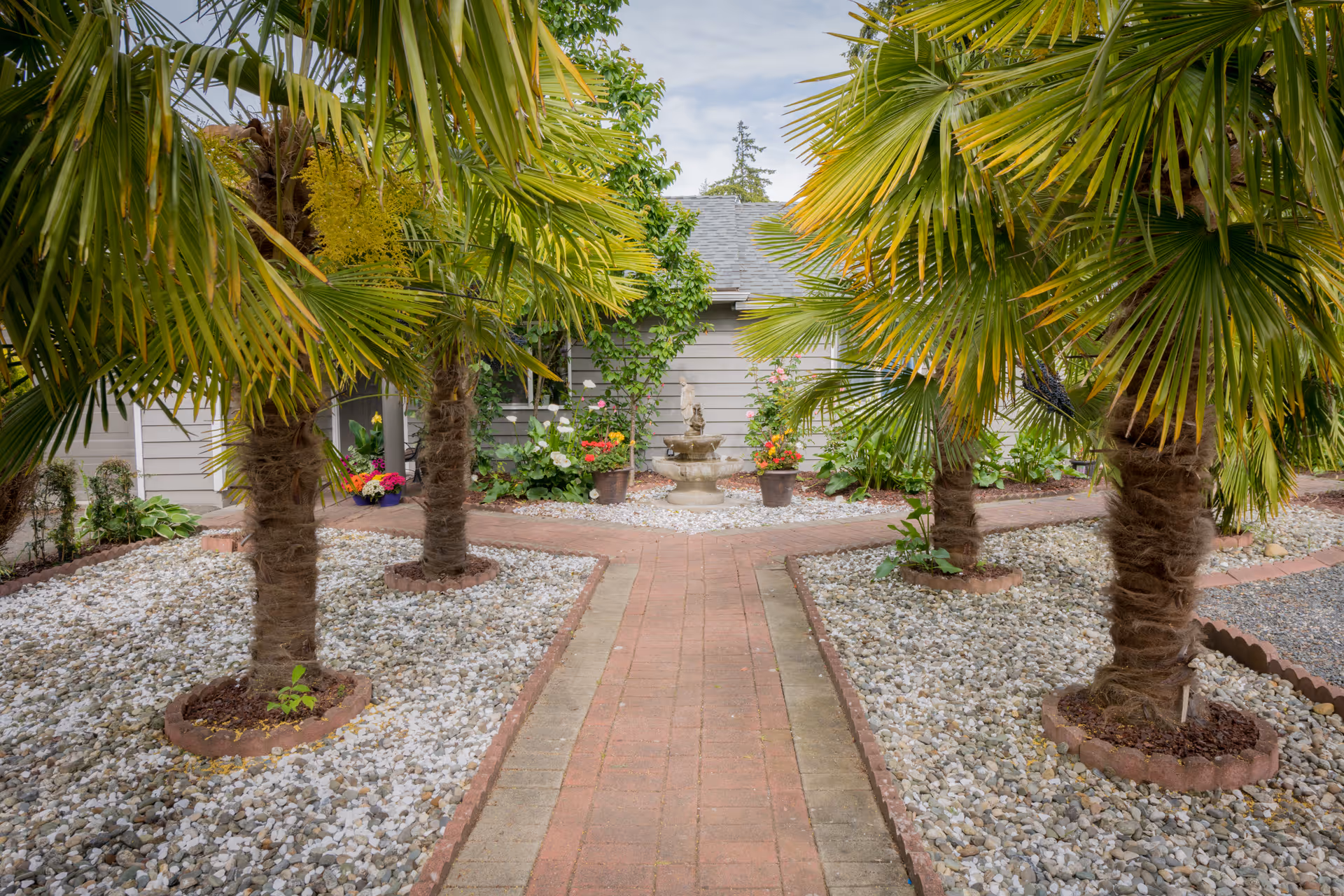 A landscaped outdoor garden area with a brick pathway leading to a small fountain surrounded by potted flowers. The garden features several palm trees planted in beds of white gravel, with a building visible in the background.