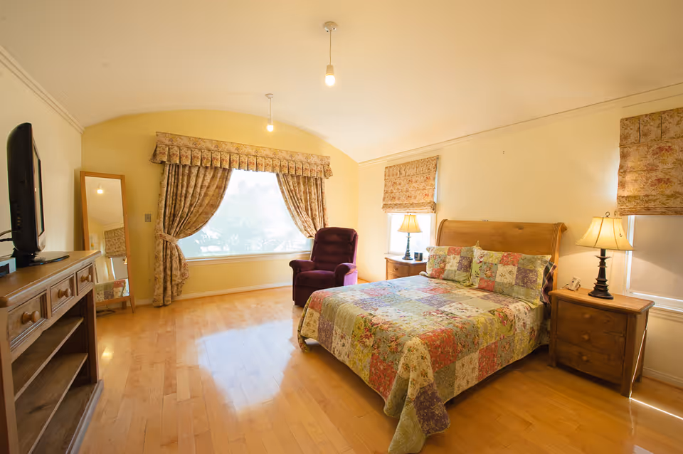 A bright bedroom with wooden flooring, a bed with a colorful patchwork quilt, two wooden nightstands with lamps, a purple armchair near a large window with floral curtains, a wooden dresser with a TV on top, and a full-length mirror leaning against the wall.