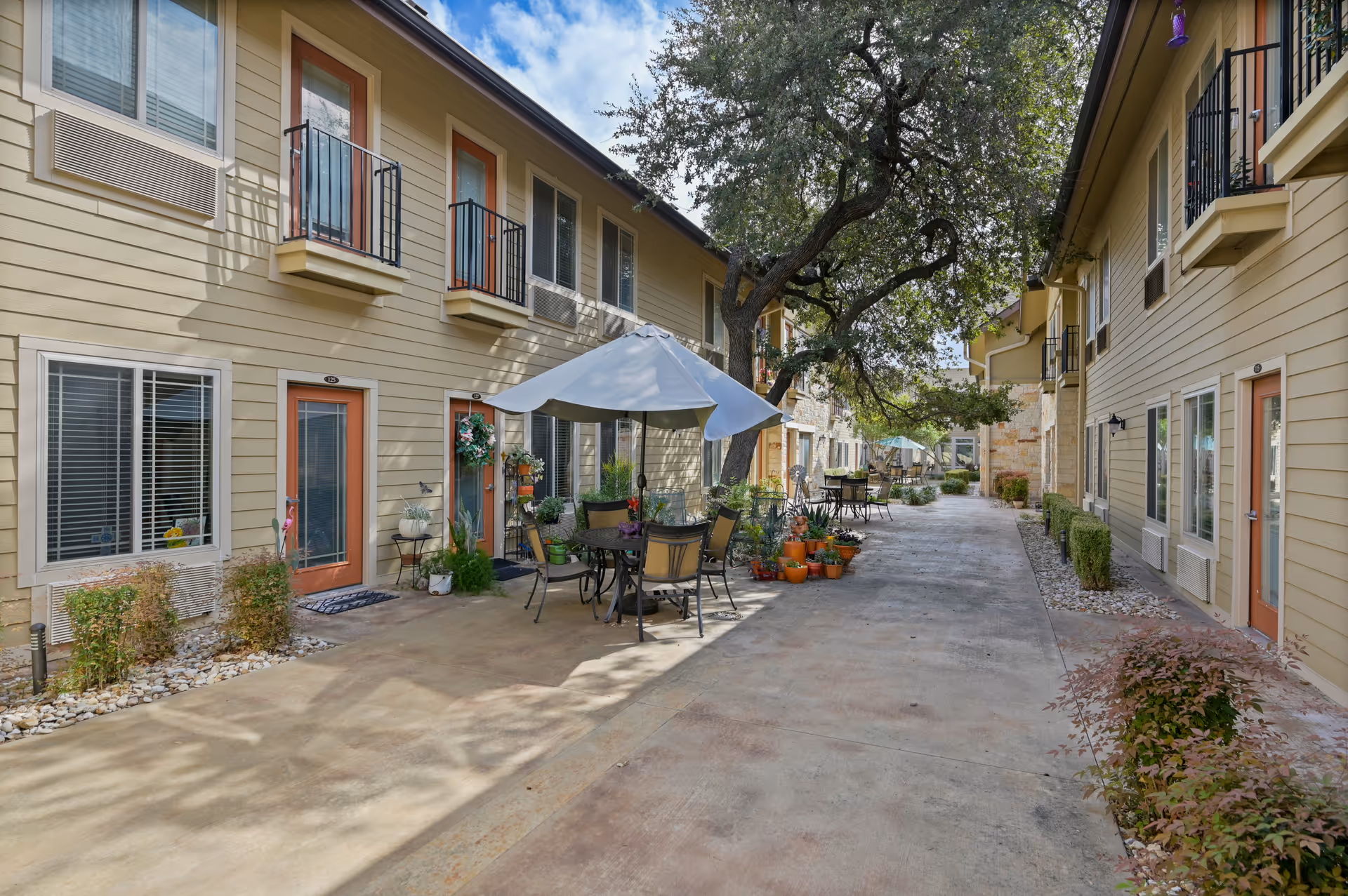 Courtyard between two-story residential buildings with outdoor tables, chairs, potted plants and a central patio umbrella.