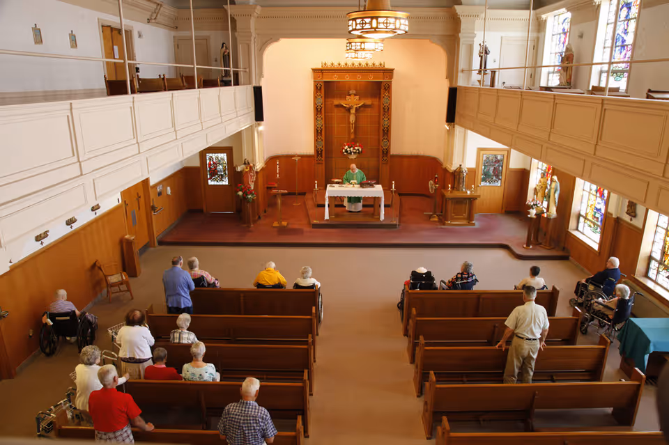 Interior view of a chapel with elderly people seated in wooden pews facing an altar where a priest is standing. The chapel has stained glass windows, religious statues, and a crucifix on the wall behind the altar.