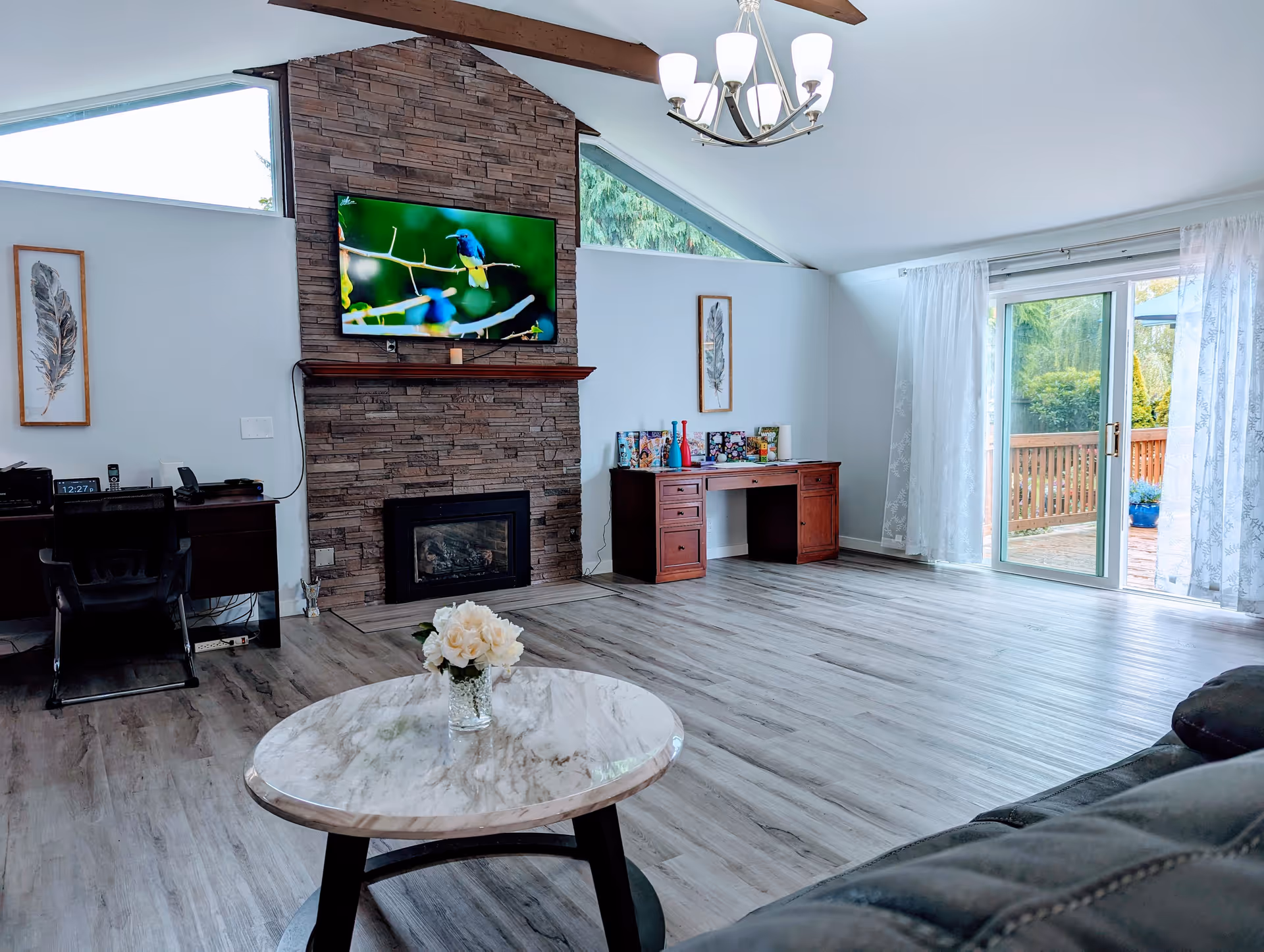 A bright living room with a stone fireplace and a mounted flat-screen TV displaying a bird. The room has light wood flooring, a round marble coffee table with a vase of white flowers, a dark leather sofa partially visible, two wooden desks with decorative items, and large sliding glass doors with sheer white curtains leading to an outdoor deck.