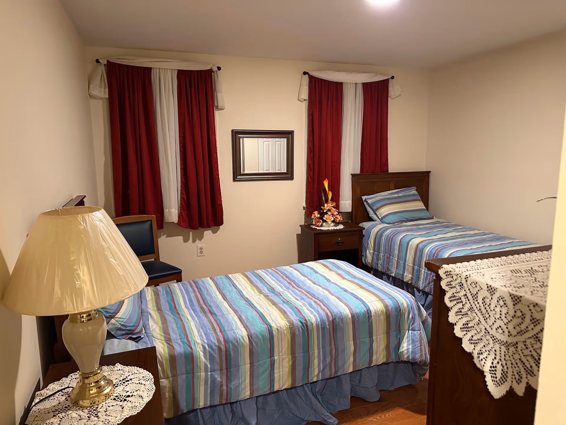 A small bedroom with two single beds covered in striped blue, green, and purple bedding. There is a wooden nightstand between the beds with a floral arrangement on top. The room has two windows with red and white curtains, a wooden chair, a table lamp with a beige shade on a small table, and a dresser with a lace doily.