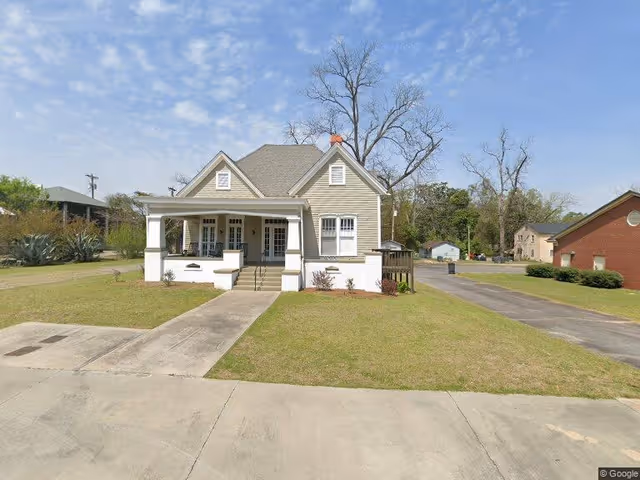 A single-story beige house with a front porch and white pillars, situated on a grassy lawn with a concrete walkway leading to the front steps. The sky is partly cloudy, and there are trees and other buildings visible in the background.
