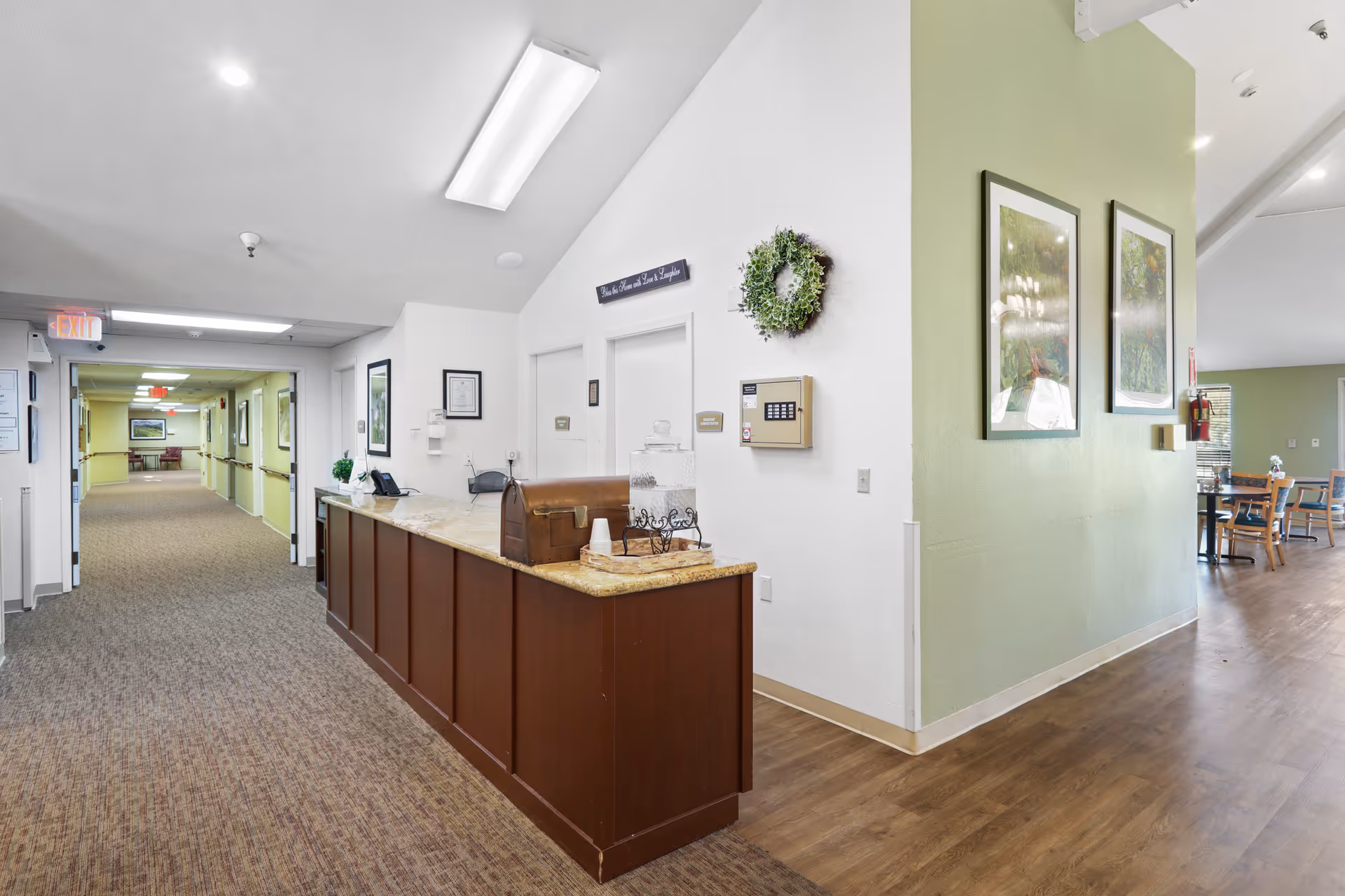 Interior view of Almond Grove Assisted Living showing a reception desk with a water dispenser and a decorative chest. The hallway extends to the left with carpeted flooring and framed pictures on the walls. To the right, there is a green accent wall with two framed pictures and a glimpse of a dining area with tables and chairs.