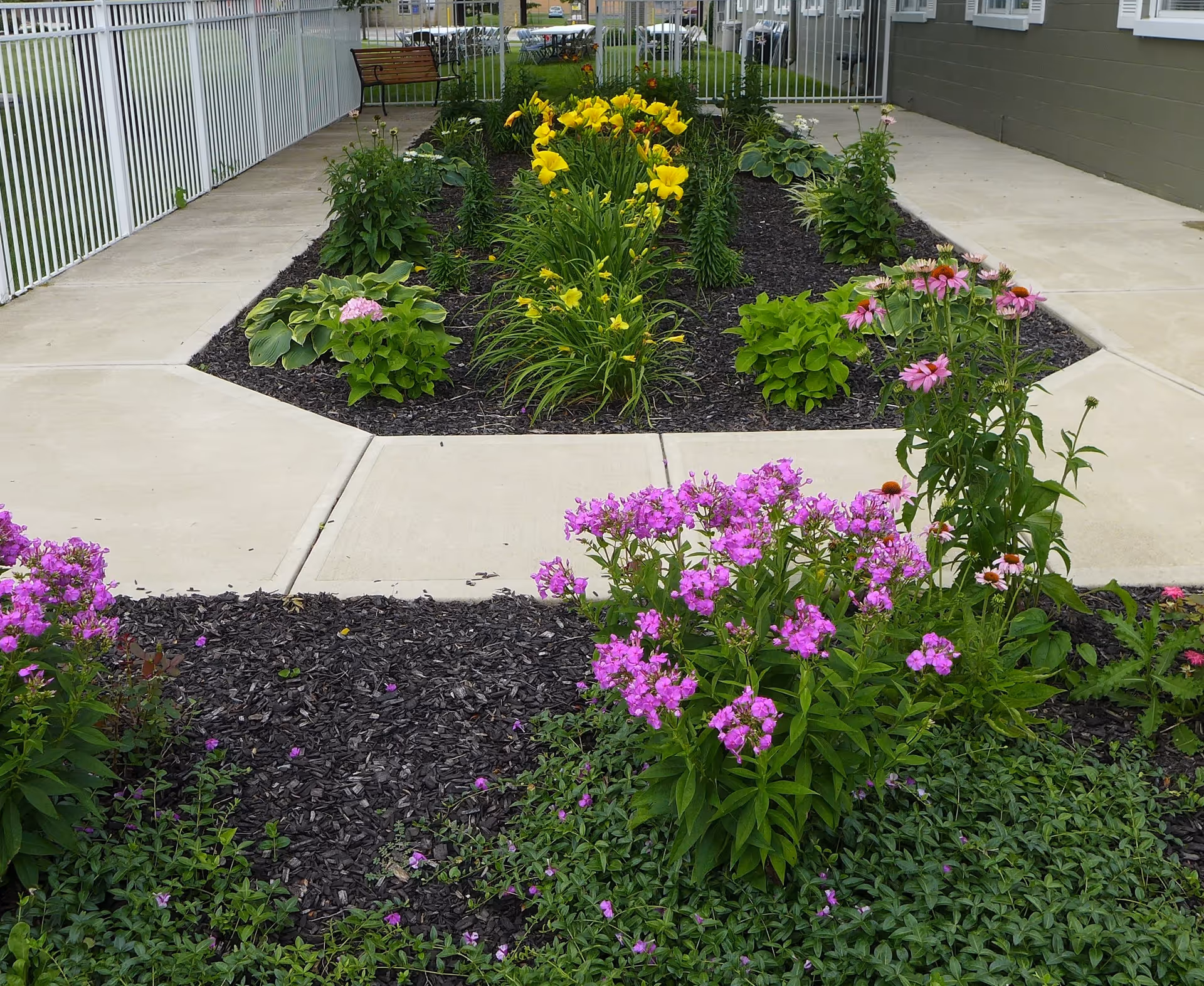 Paved courtyard garden with beds of pink and yellow flowers, a white fence, and a bench beside a building.