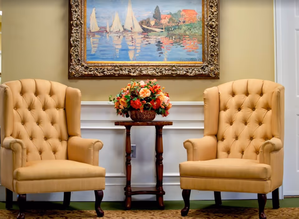 Two beige tufted armchairs with wooden legs are positioned on either side of a small wooden table holding a floral arrangement with red, orange, and pink flowers. Above the table hangs a framed painting depicting sailboats on water with reflections and buildings in the background. The setting appears to be a cozy, well-decorated interior space with light-colored walls and a green carpet.