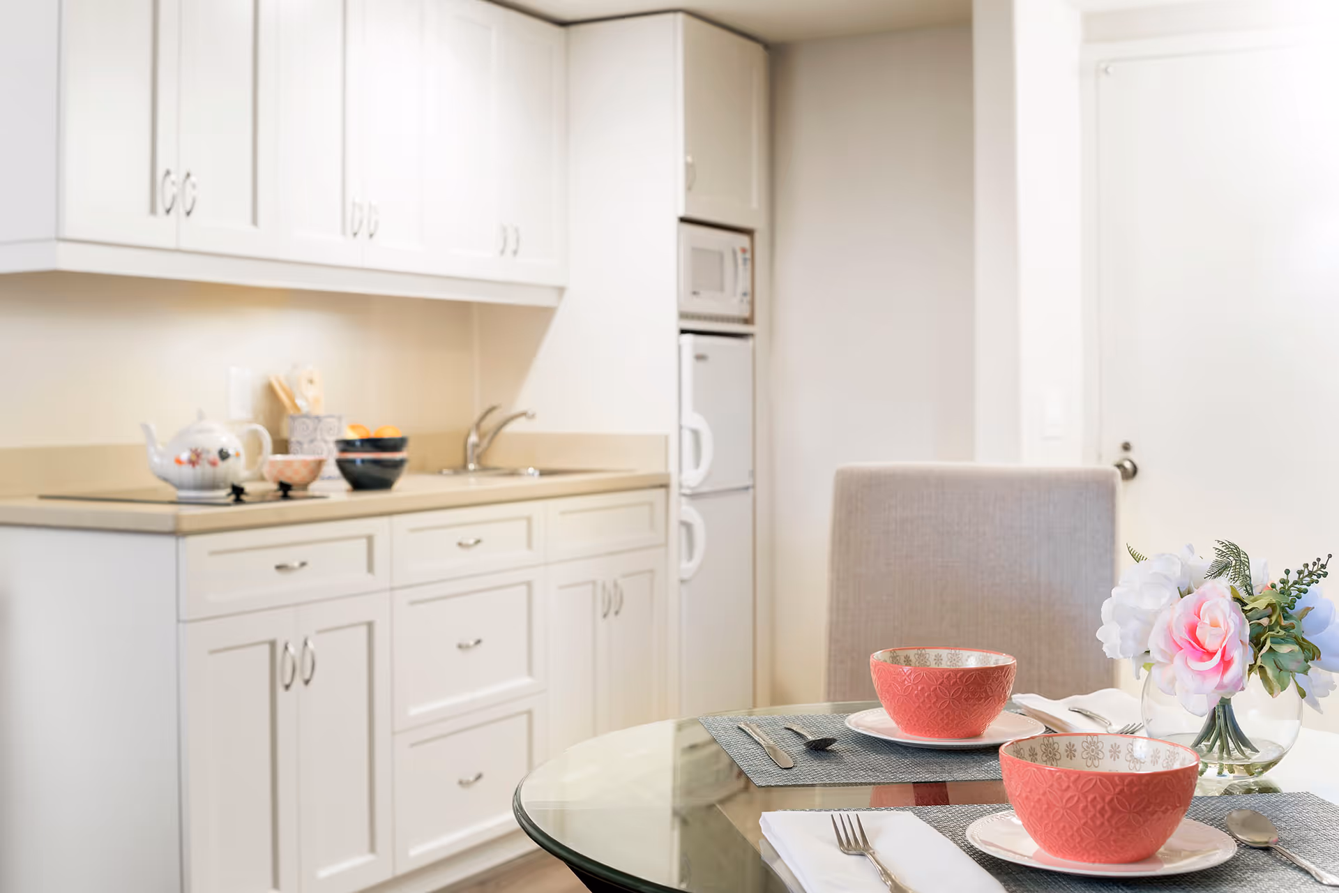 A bright kitchen area with white cabinets, a countertop with a teapot, bowls, and utensils. In the foreground, a glass dining table is set with two red patterned bowls on white plates, silverware, and a small vase with pink and white flowers. A beige upholstered chair is visible behind the table.