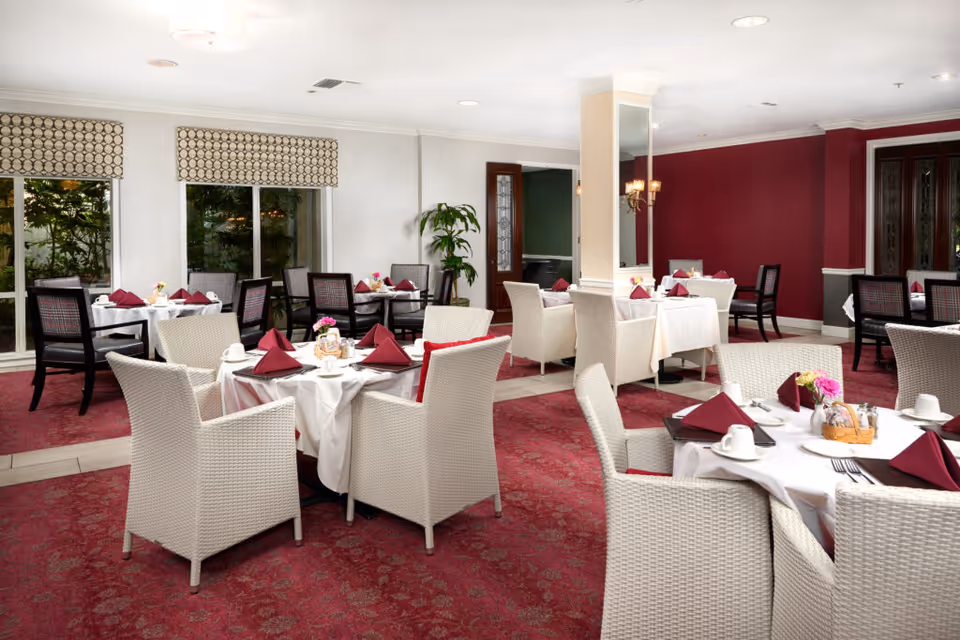 Dining room with multiple round tables set with white tablecloths, burgundy napkins, wicker and upholstered chairs, and a red patterned carpet.