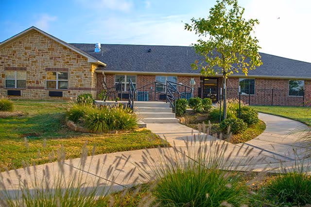 Single-story building with a combination of stone and brick exterior walls, a gray shingled roof, and several windows. A concrete pathway leads to the entrance with steps and black metal railings. The surrounding area has green grass, bushes, and a tree under a clear blue sky.