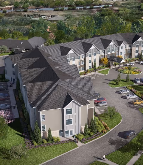 Aerial view of a large senior living facility building with multiple gabled roofs, surrounded by landscaped greenery and parking areas with several cars. The facility has a driveway leading to a covered entrance and is set in a suburban area with trees and other buildings in the background.