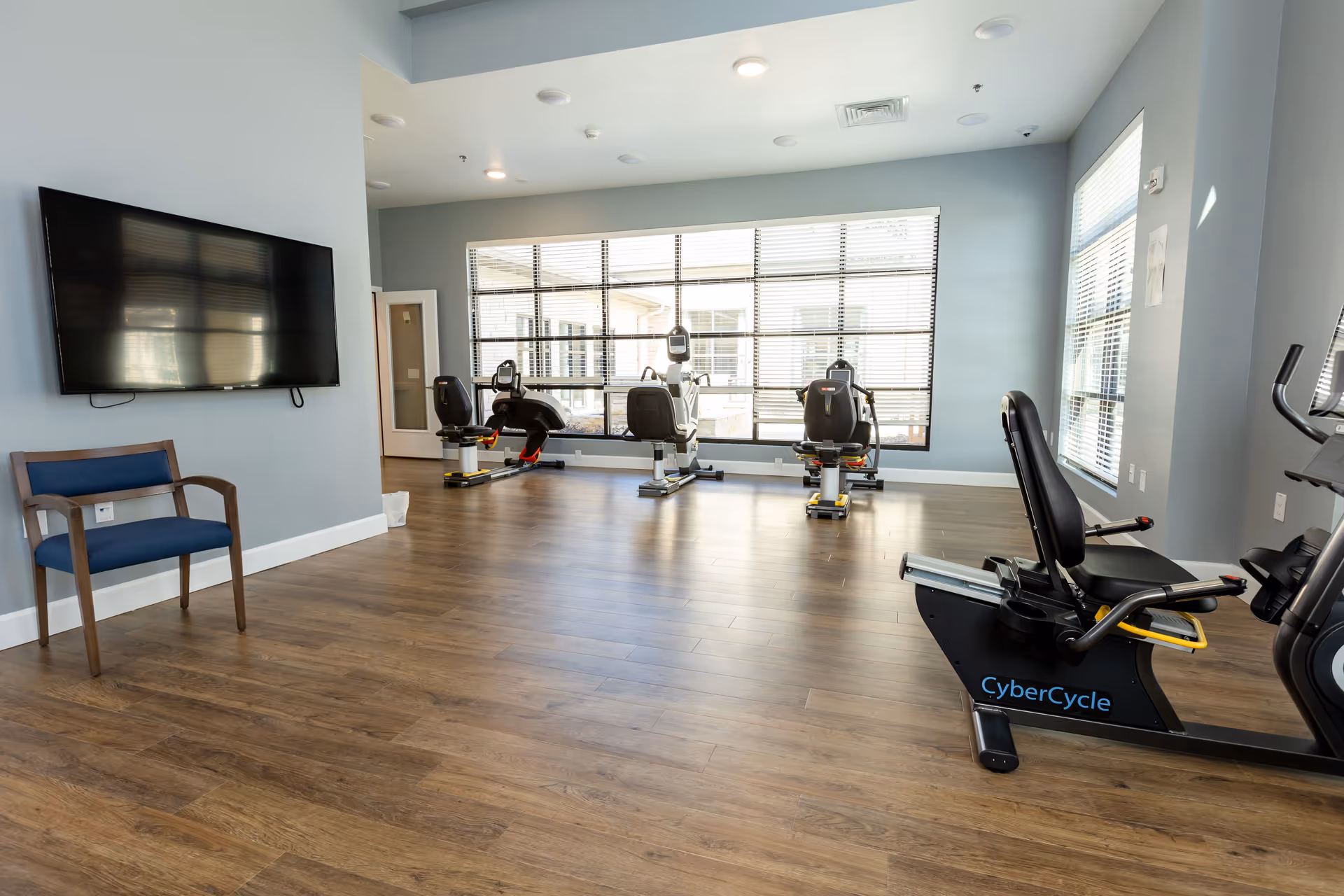 A bright exercise room with wooden flooring and light blue walls. The room contains several recumbent exercise bikes positioned near large windows with blinds. A flat-screen TV is mounted on one wall, and a blue cushioned wooden chair is placed beneath it.