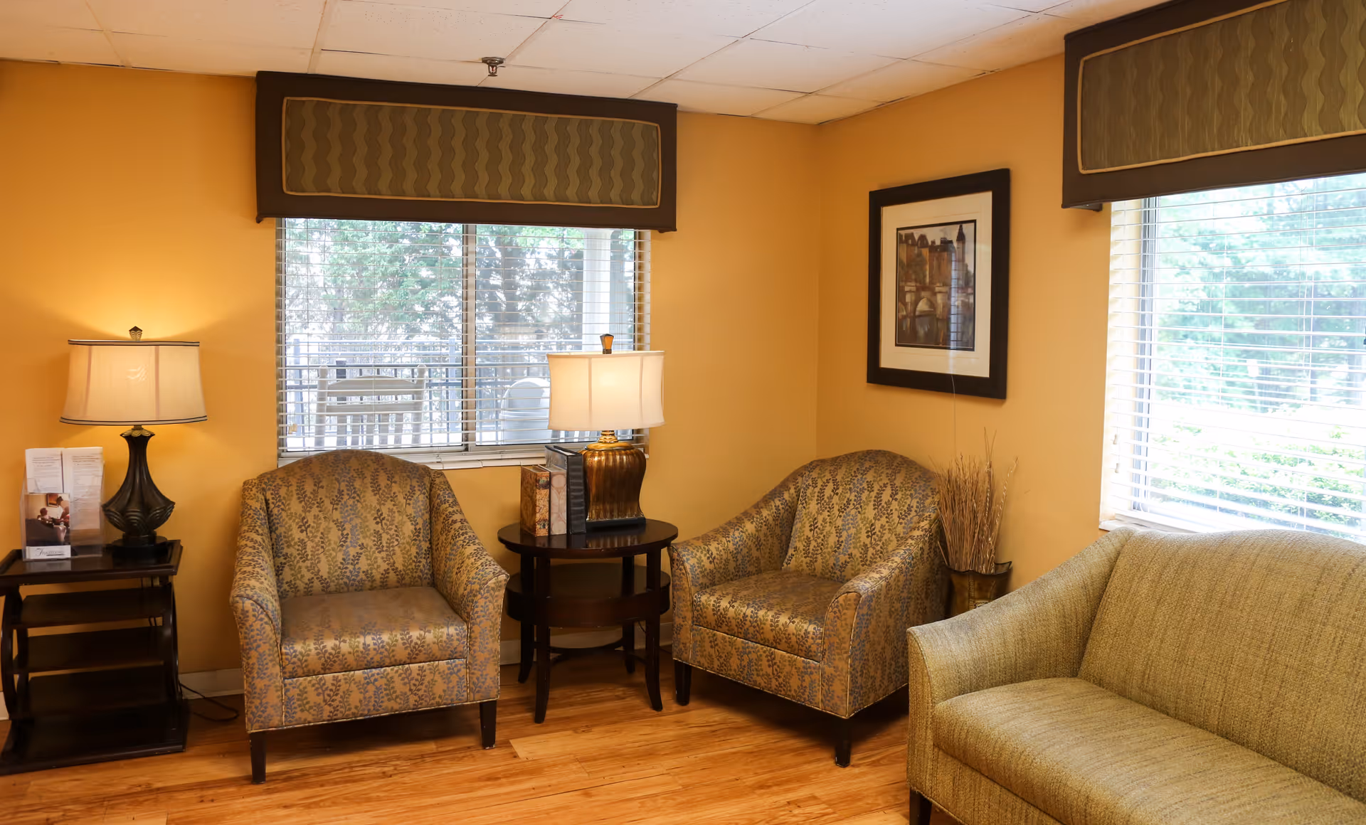 A cozy sitting area in a senior living facility with two patterned armchairs and a green sofa arranged around two wooden side tables with lamps. The walls are painted warm yellow, and there are two windows with blinds letting in natural light. A framed picture and a decorative vase with dried plants are on the walls and floor.