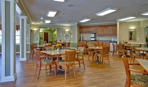 A spacious dining room with multiple wooden tables and chairs arranged neatly on a wood floor. The room features soft lighting from ceiling fixtures and wall sconces, with a kitchen area in the background equipped with cabinets, a microwave, and a coffee machine. The walls are decorated with framed artwork and mirrors, creating a warm and inviting atmosphere.