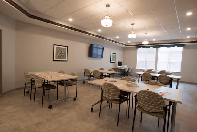 Well-lit communal dining/activity room with multiple tables and patterned chairs, a wall-mounted TV, and large windows.