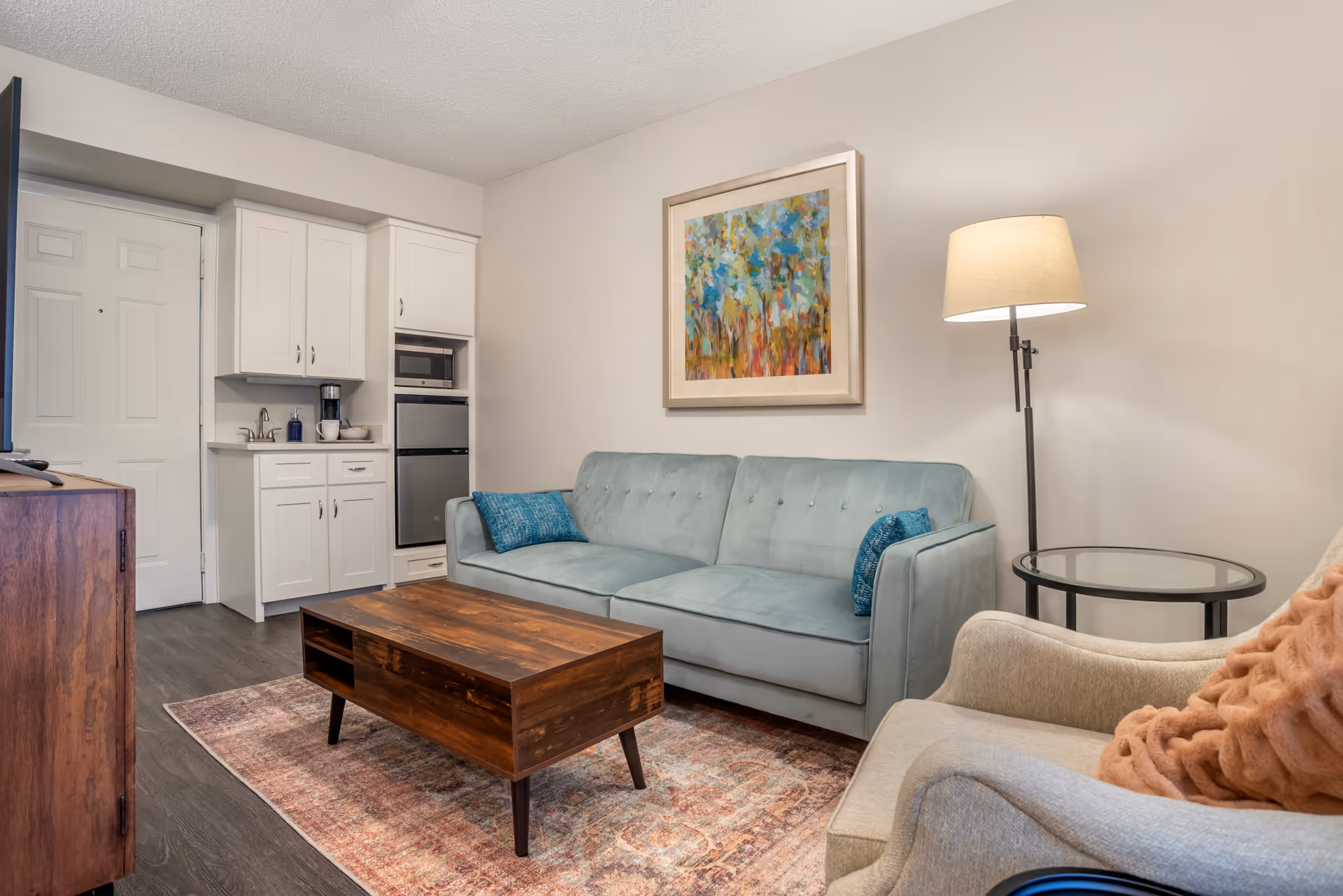 Small living room with a teal sofa, wooden coffee table, armchair, floor lamp, wall art, and a compact kitchenette in the corner.