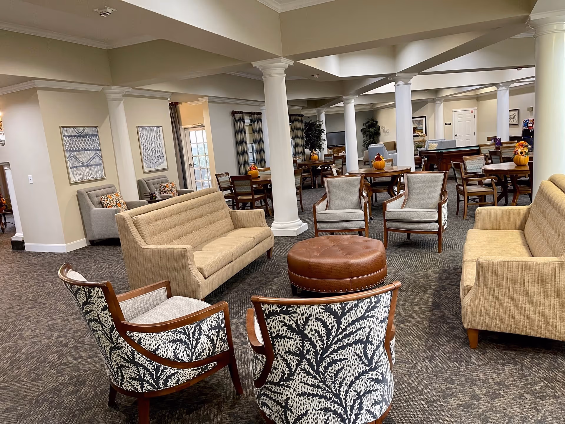 A spacious assisted living facility common area with multiple seating arrangements including beige sofas, patterned armchairs, and wooden chairs around round tables. The room features white columns, carpeted floors, and neutral-colored walls with framed artwork. There are decorative pumpkins on the tables, and the area appears well-lit with natural light coming through windows.