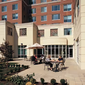 Paved courtyard with round patio tables, umbrellas, and people seated outside a multi-story brick-and-beige residential building.