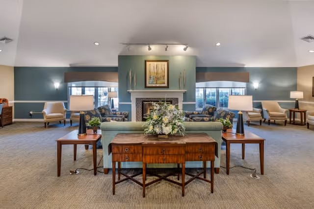 A spacious and well-lit living room area in a senior living facility featuring a central fireplace with a framed painting above it. The room has multiple seating arrangements including armchairs and sofas, side tables with lamps, and decorative plants. Large windows with valances allow natural light to fill the space.
