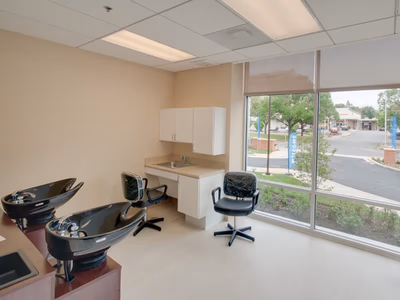 Bright salon-style room with two hair-washing stations, several swivel chairs, a sink with cabinets, and large windows overlooking the parking lot.