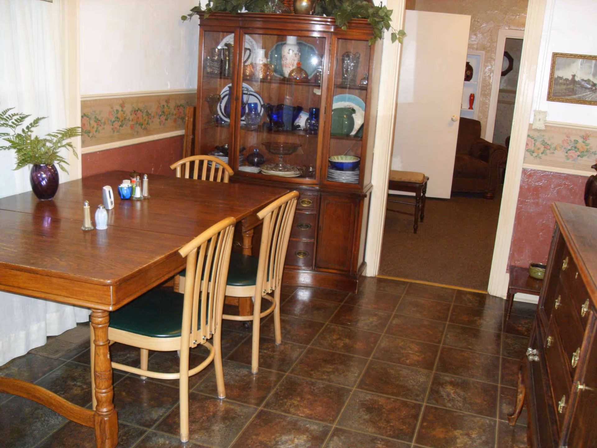 A dining room with a wooden table and four chairs, a glass-fronted china cabinet filled with dishes and decorative items, floral wallpaper border, and a doorway leading to another room with a brown armchair and a small bench.