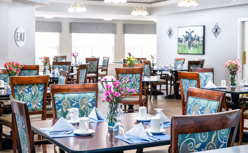 A bright dining room with multiple wooden tables and chairs featuring blue patterned upholstery. Each table is set with white cups, glasses, folded napkins, and silverware. Vases with pink flowers decorate the tables. Large windows with gray valances allow natural light to fill the room. Wall decor includes a round sign with the word 'EAT' and a framed picture of cows in a field.