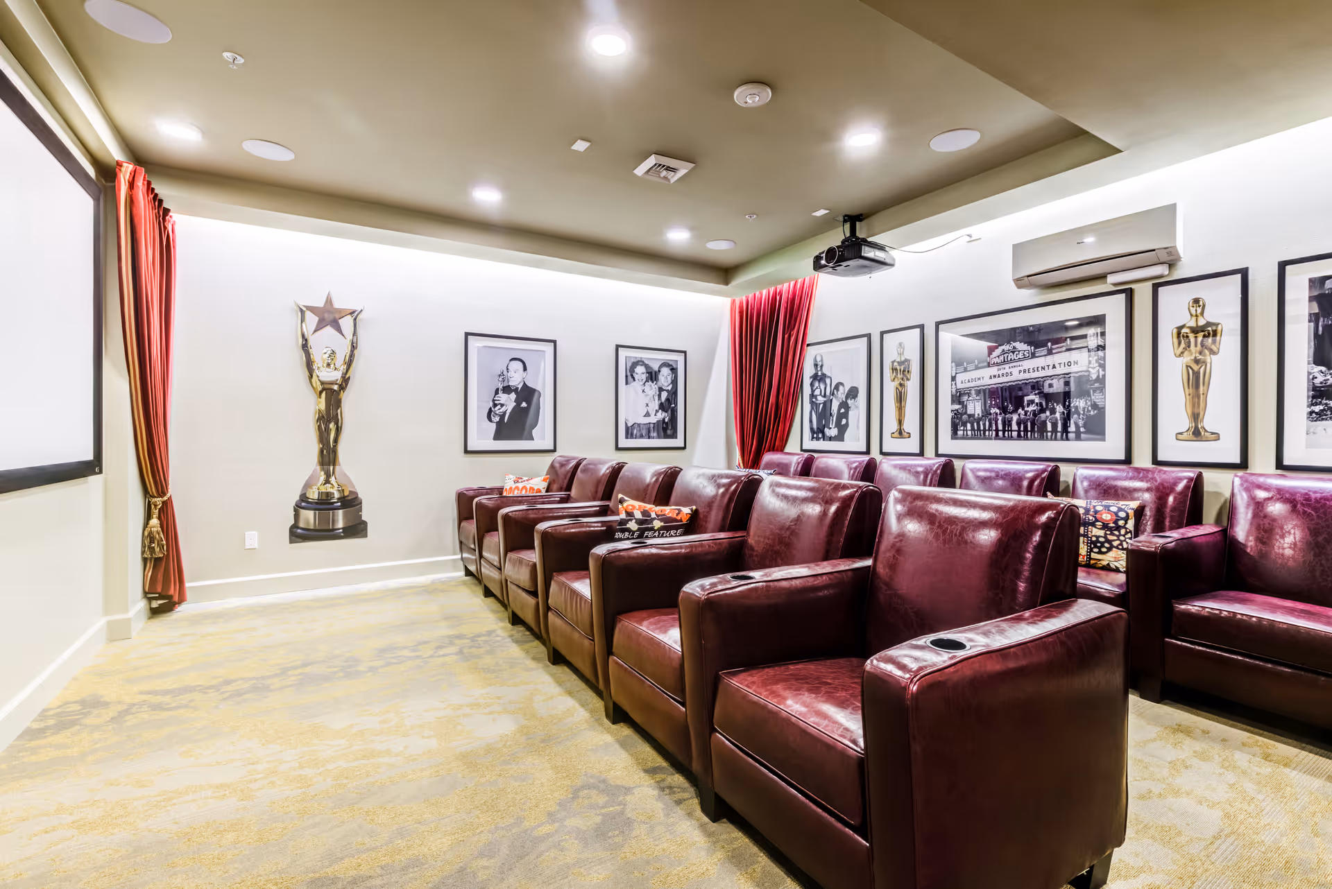A small home theater room with two rows of burgundy leather recliner chairs facing a large projection screen. The walls are decorated with framed black and white photos and images of Oscar statues. Red curtains frame the screen area, and a projector is mounted on the ceiling.