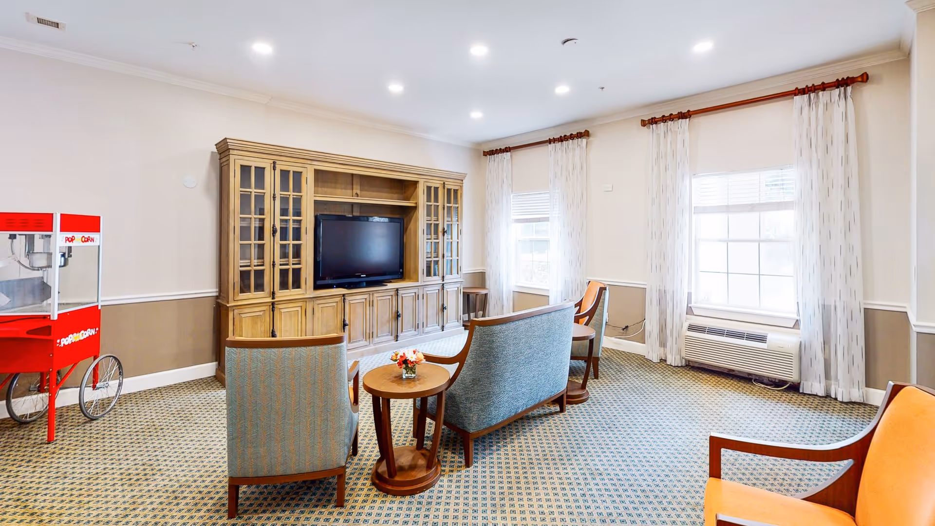 A cozy living room area with a wooden entertainment center holding a flat-screen TV, surrounded by upholstered chairs and a loveseat. There is a small round wooden table with a flower arrangement in the center. Two large windows with sheer white curtains allow natural light to fill the room. A red popcorn machine is positioned in the corner.