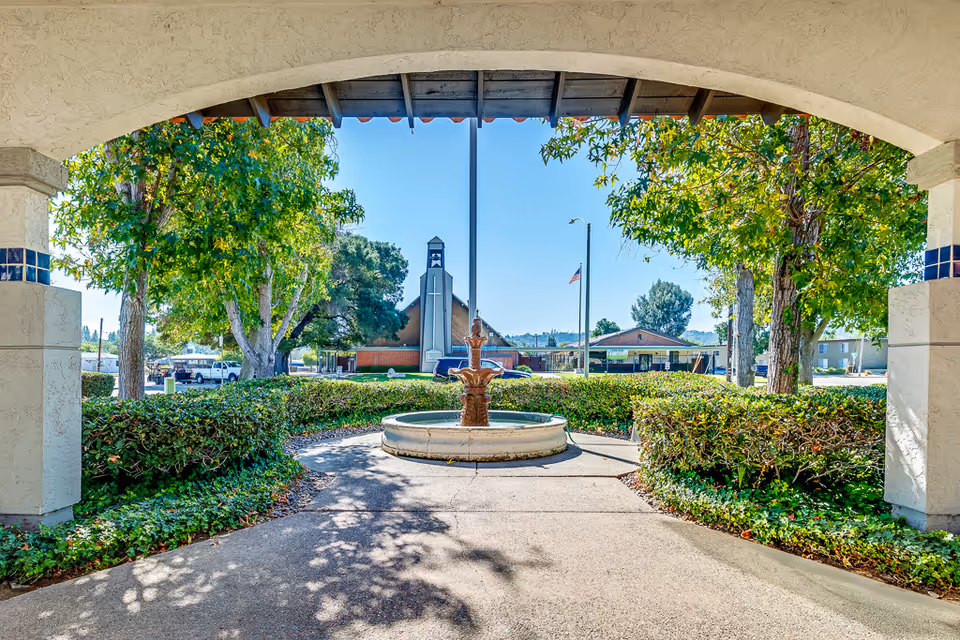 View through an arched entrance showing a circular fountain surrounded by green bushes and trees. In the background, there is a building with a tall structure featuring a cross, an American flag on a pole, and a clear blue sky.